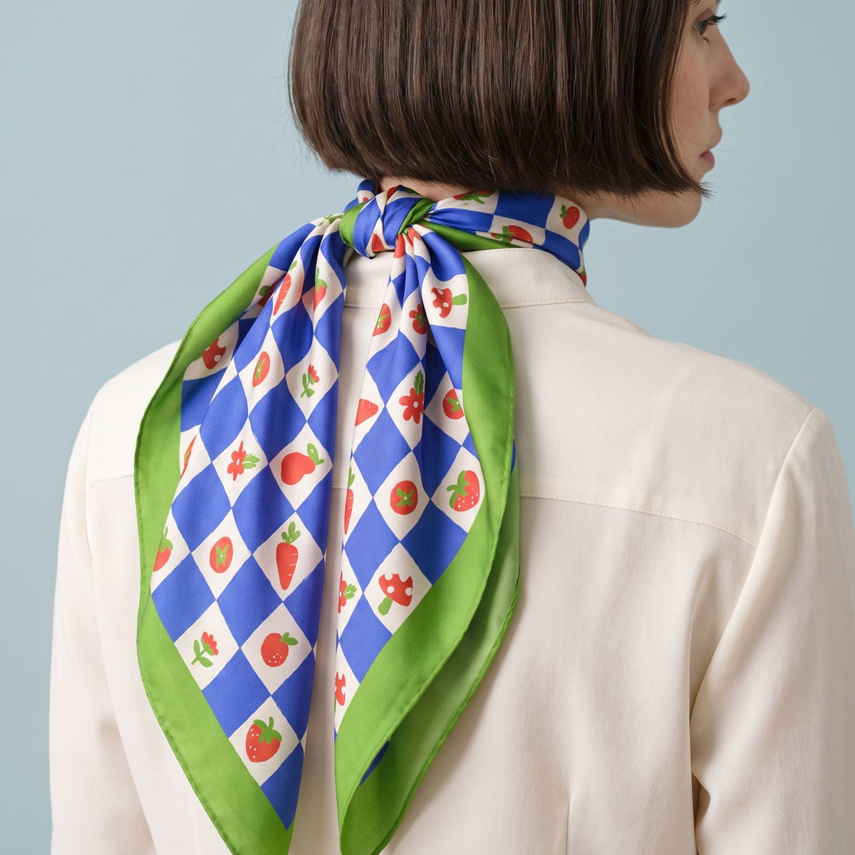 A woman shown from the back wearing a cream-colored blazer and a checkers patterned silk scarf tied around her neck. The scarf features a blue and white argyle design with red strawberries and green leaves, finished with a solid green border, set against a light blue background.