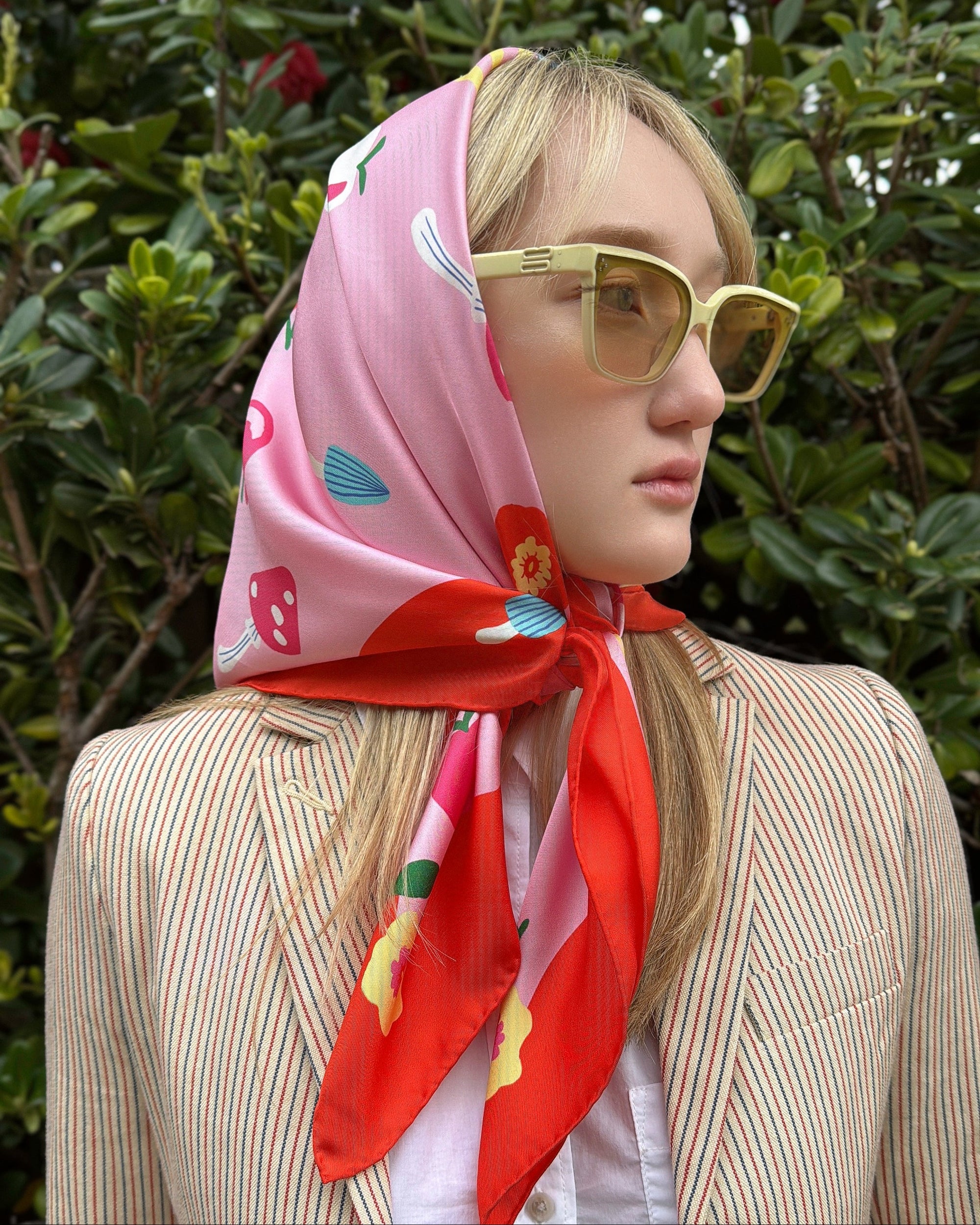 Person wearing a colorful cat garden gnome silk scarf on pink and red with flowers and mushrrooms and sunglasses with a blurred green background 