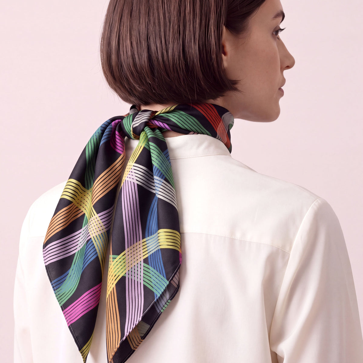 A woman with a dark brown bob haircut is shown from the back and side profile, wearing a white collared shirt and a colorful striped silk scarf tied around her neck, against a solid pale pink background.