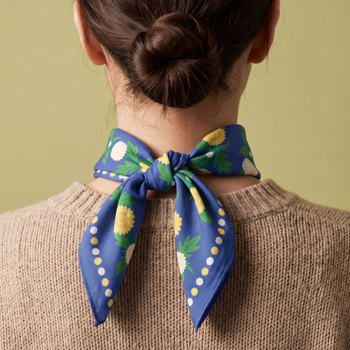 A person viewed from the back wearing a brown knit sweater and a blue dandelion patterned bandana cotton scarf tied in a bow, against a solid olive-green background.