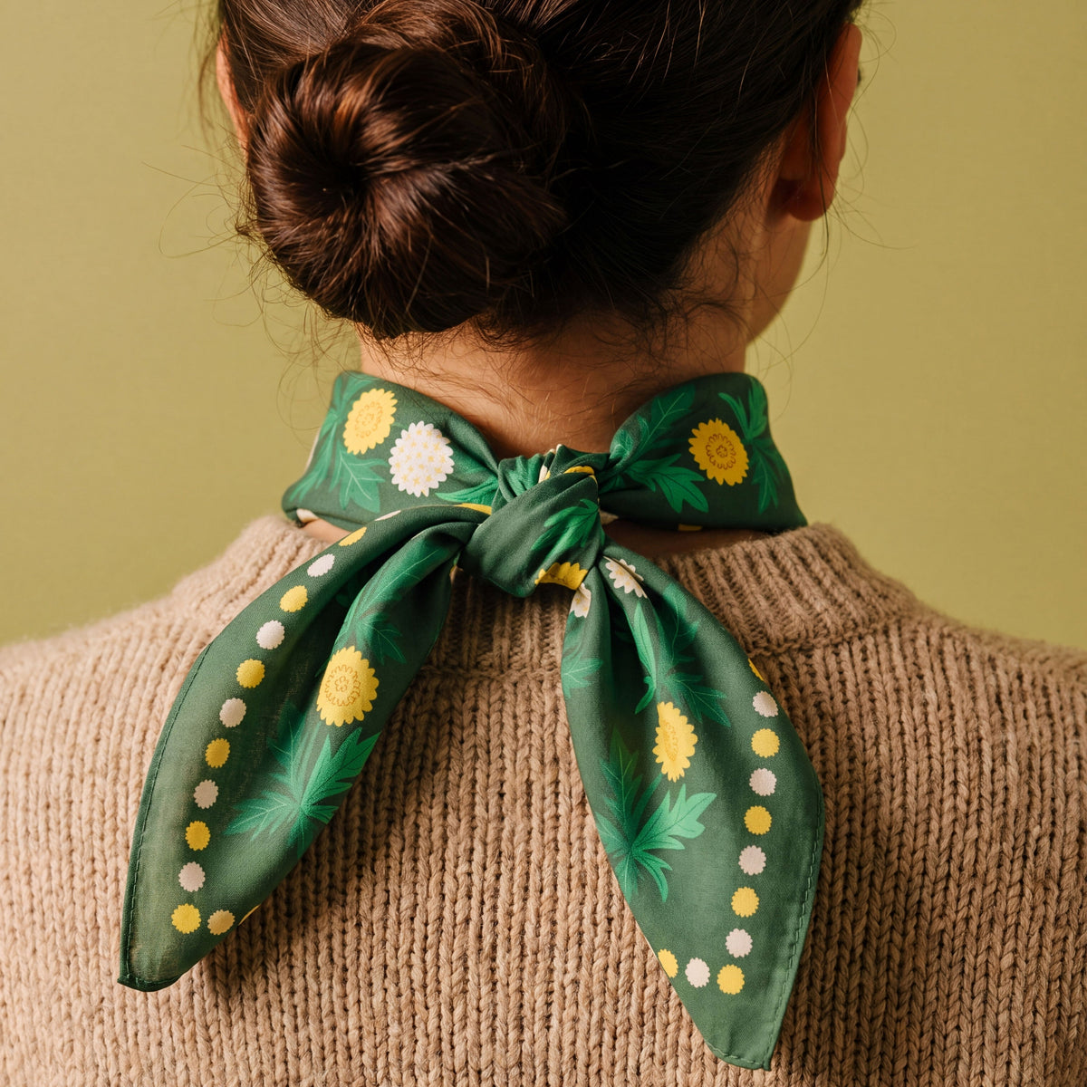 A woman viewed from behind wearing a green cotton bandana tied around her neck. The scarf features a pattern of yellow dandelion and white daisies floral with yellow and white dots along the edges.