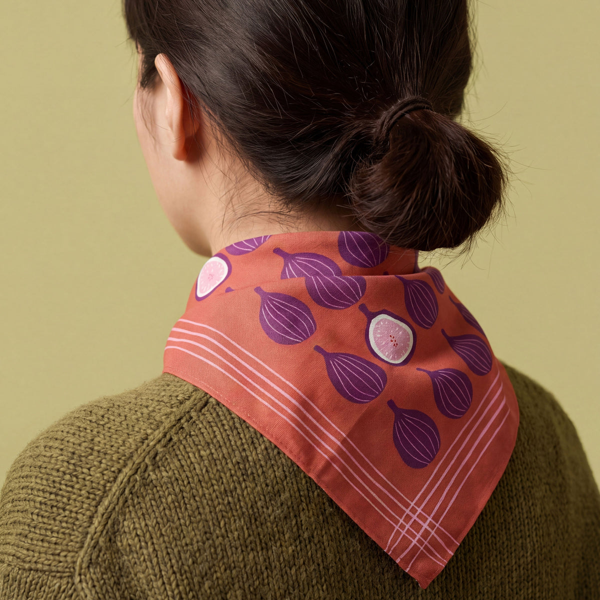 A person shown from the back wearing a terracotta orange bandana cotton scarf with a purple and white fig pattern, styled over an olive green knit sweater against a solid beige background.