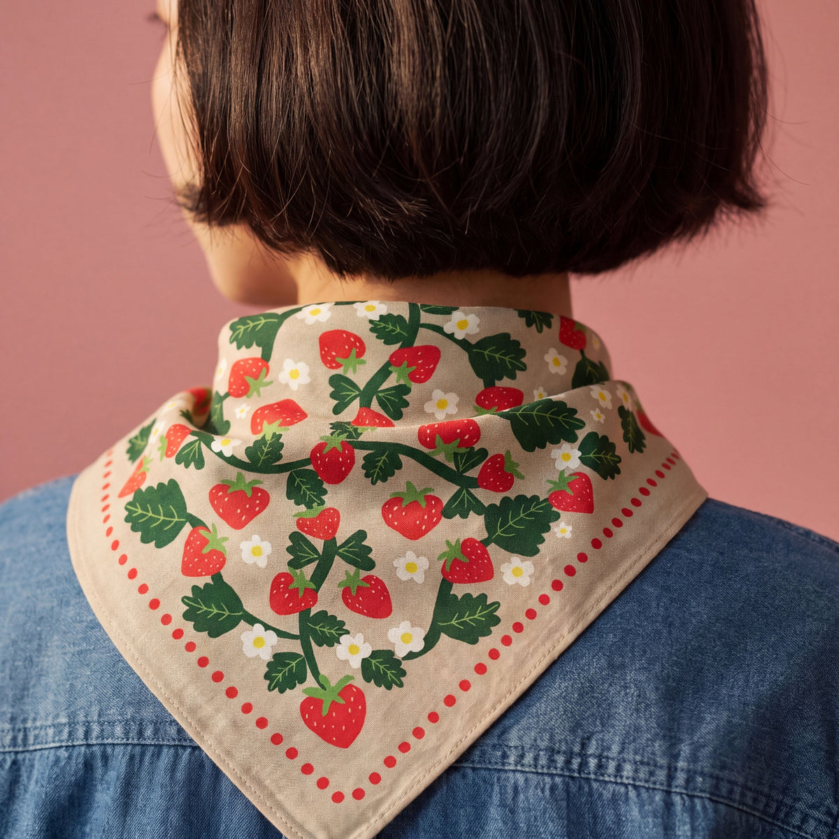 A close-up back view of a person with short dark hair wearing a beige neck scarf printed with red strawberries, green leaves, and small white flowers, over a blue denim shirt against a dusty pink background.
