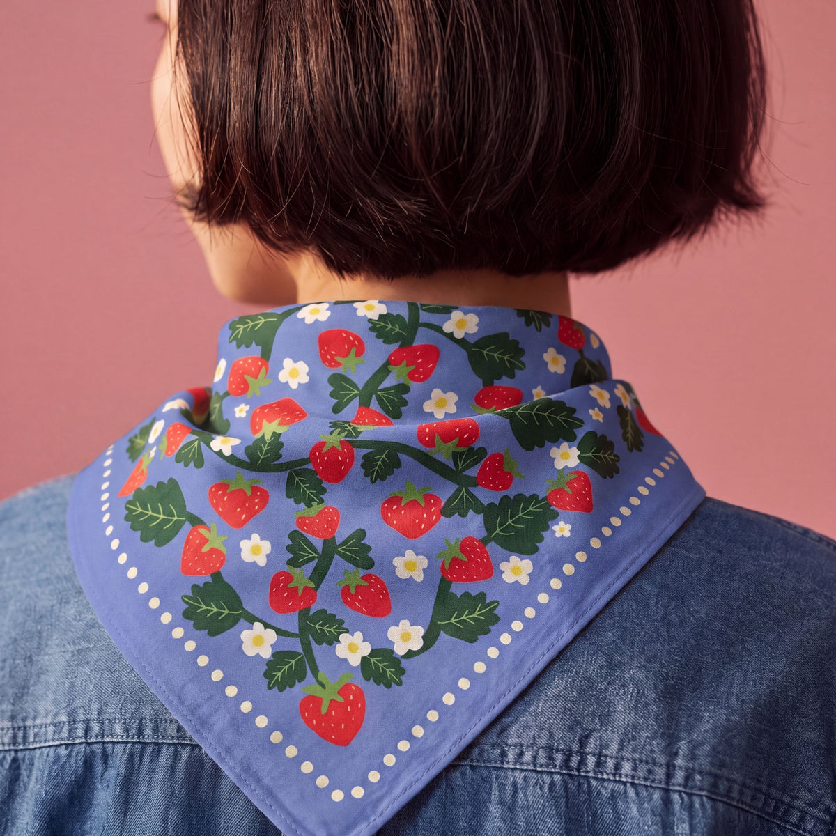 A close-up back view of a person with a short dark bob haircut, wearing a blue denim shirt and a light blue bandana-style neck scarf printed with red strawberries, green leaves, and small white flowers against a solid pink background.