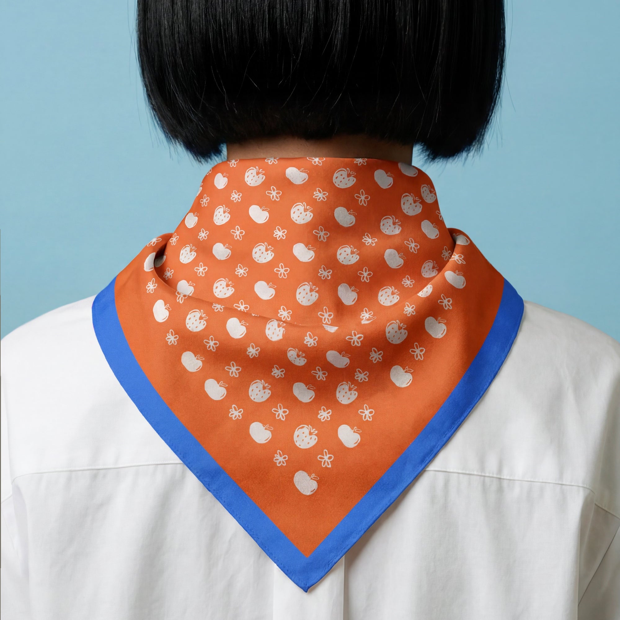 woman wearing an orange scarf with white apples, strawberries and flowers pattern, with blue border on a blue background